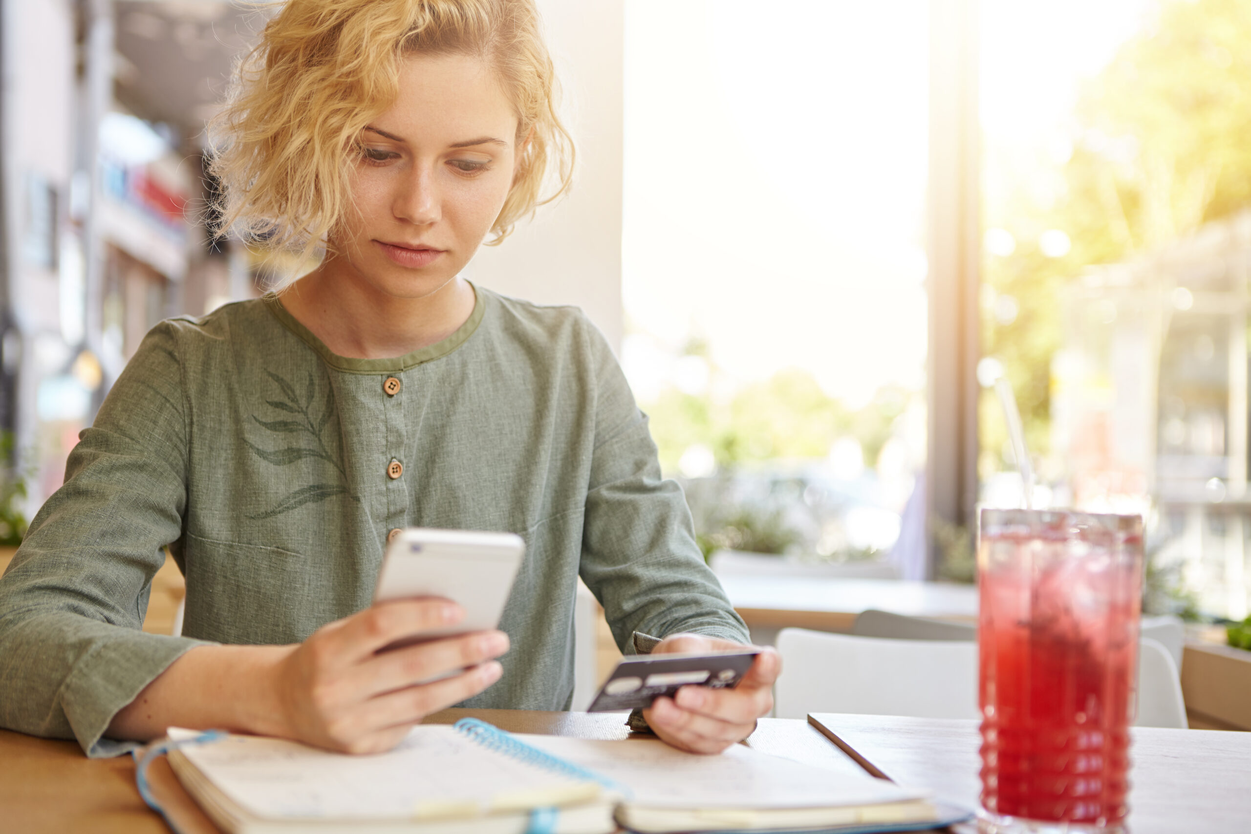 Elegant blonde woman in blouse sitting at cafe holding mobile phone and plastic card signing up on website. Businesswoman with stylish hairdo paying with credit card while shopping online using mobile