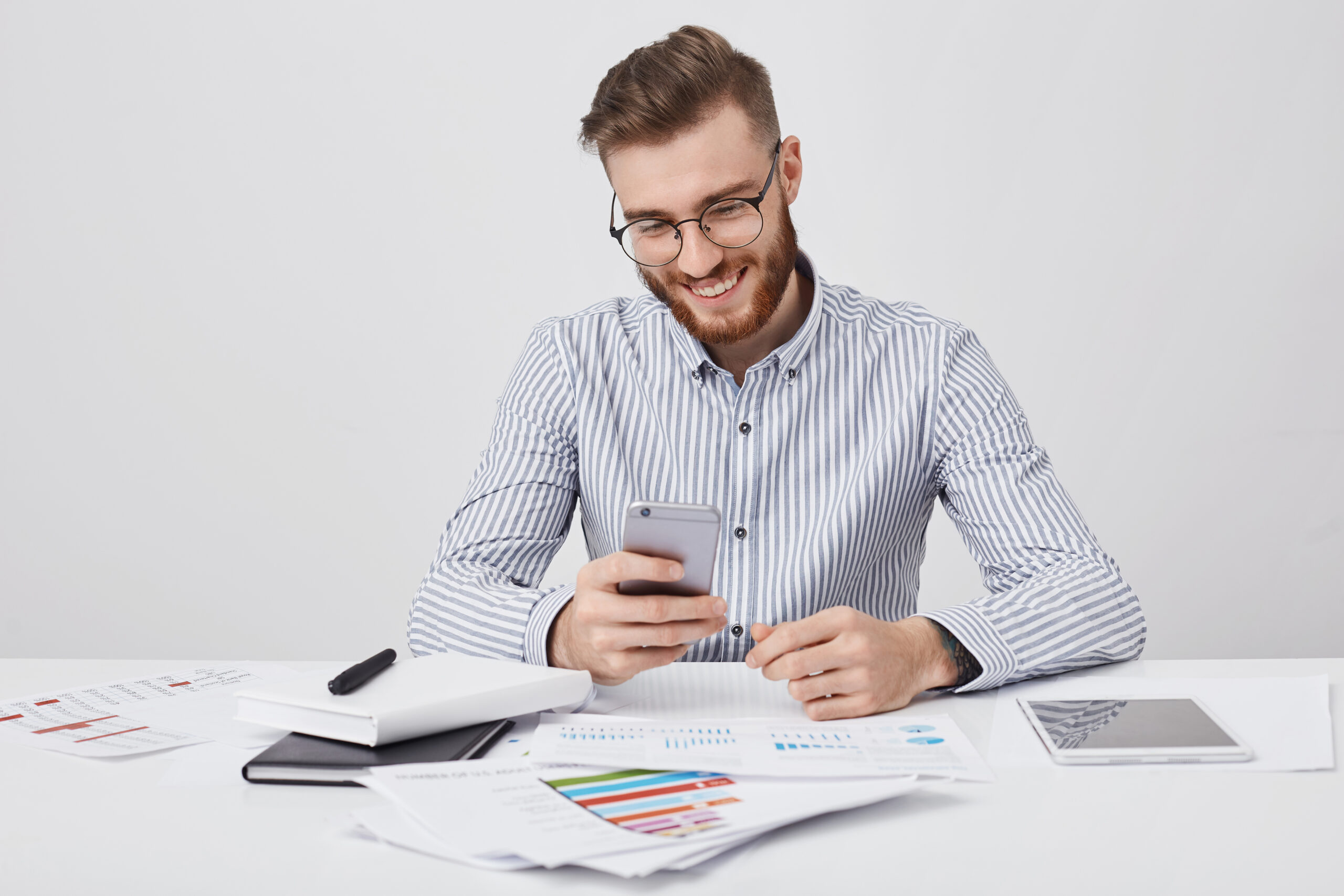 People, technology, occupation and online communication concept. Smiling male manager surrounded with many papers and gadgets, recieves congratulations on cell phone from friend as has birthday